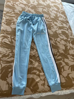 Palm Angels ljusblå mjukisbyxor - Palm Angels mjukisbyxor i ljusblått med vita och svarta sidoremsor. Byxorna har elastisk midja med snörning och ribbade muddar vid bensluten. Märkets logga är tryckt framtill. Materialet är mjukt och syntetiskt, perfekt för chill eller träning.