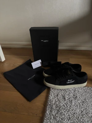 Svarta Saint Laurent skor.  - Svarta sneakers från Saint Laurent med vit logotyp på sidan. Skorna har snörning, rund tå och en beige platt sula. Använda 1 gång för de va för små. Tveka inte om du har frågor eller vill ha fler bilder☺️Byten kan va intressanta.