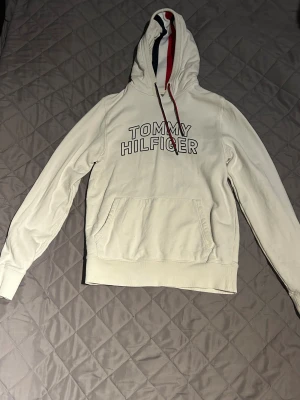 Vit hoodie Tommy Hilfiger - Vit hoodie från Tommy Hilfiger med stor logotyp på bröstet och klassiska röd-blå detaljer i huvan. Tröjan har snörning vid huvan, magficka och långa ärmar. Mjuk bomullskänsla och clean look, perfekt för chill dagar.