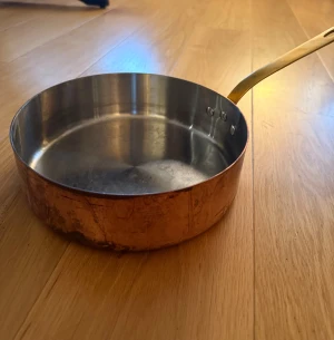 Koppargryta med mässingshandtag - Robust koppargryta med insida i rostfritt stål och mässingshandtag. Grytan har tydliga tecken på användning, inklusive repor och patina på kopparytan, men är i gott bruksskick. Perfekt för matlagning med jämn värmefördelning. Ingen synlig allvarlig skada. Diameter 21cm