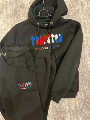 Trapstar svart set med broderi - Svart set från Trapstar med broderad logga i blått, rött och vitt på bröstet. Tröjan har huva med snörning, ribbade muddar och känguruficka. Materialet är mjukt och känns som bomull eller polyester, perfekt för chill eller streetwear. Snygg och unik design med färgstarka detaljer.