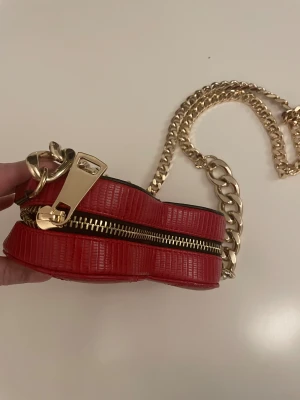 Röd hjärtformad axelväska River Island - Säljer en röd axelväska från River Island i hjärtformad design med guldfärgad kedja och detaljer. Väskan är tillverkad i skinnimitation och har quiltade sömmar samt en snygg dragkedja. Perfekt för dig som vill sticka ut med en unik accessoar.18cmx6cm . Aldrig använt som nytt !