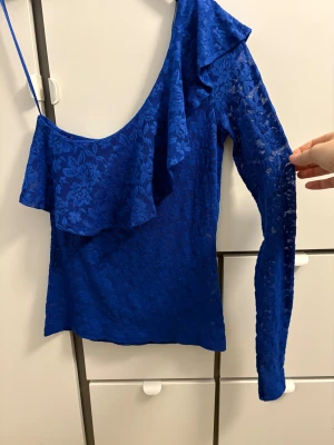 Blå spetsig oneshoulder topp Gina Tricot - Säljer en blå oneshoulder topp från Gina Tricot i storlek S. Toppen har lång ärm på ena sidan, volangdetalj och är gjord i blommig spets. Snygg och unik design med smal passform och transparent känsla. Perfekt för dig som vill sticka ut med färg och form.