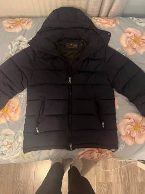 Svart pufferjacka från Oscar Jacobson - Säljer en svart pufferjacka från Oscar Jacobson med huva och tvåvägsdragkedja. Jackan är vattenavvisande och isolerande, perfekt för kalla dagar. Tillverkad i syntetmaterial med Sorona/Dupont-fyllning. Snygg och stilren design med horisontella sömmar och praktiska fickor med dragkedja. Jag har använt den typ 5 gånger och den kostar 6000kr från butiken.