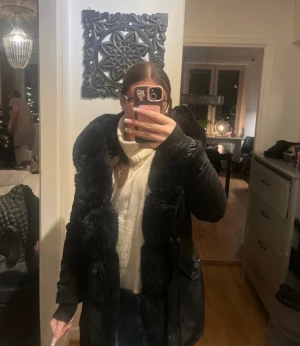 Svart parkas med stor fuskpäls - Säljer en svart parkas med stor fluffig fuskpäls runt huvan och längs öppningen. Jackan har dragkedja, knappar och flera fickor framtill. Snygg och varm modell med justerbar midja och långa ärmar. Perfekt för kalla dagar och riktigt mysig att bära.