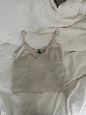 Champagnefärgad satin linnetopp DIVIDED - Säljer en champagnefärgad linnetopp från DIVIDED i satin med spetsdetaljer och liten rosett framtill. Toppens smala axelband och glansiga material ger en lyxig vibe. Perfekt för dig som gillar stilrena och romantiska plagg.