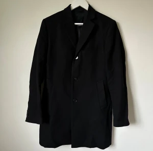 Svart rock från J.Lindeberg, stl 46 - Stilren svart rock från J.Lindeberg i klassisk modell med knappar framtill och fickor på sidorna. Tillverkad i 80% ull och 20% polyamid med foder i 100% viskos. Perfekt för dig som vill ha en clean och tidlös look. Modell: Wolger 77 Compact Melton - 4C6 