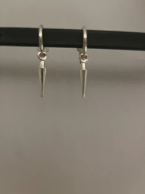 Silvriga örhängen med spetsig hängande detalj - Säljer ett par snygga  örhängen i silverfärg med hängande spetsiga detaljer. De har en minimalistisk och edgy design som ger en modern vibe. Perfekta för dig som gillar stilrena smycken med en twist.