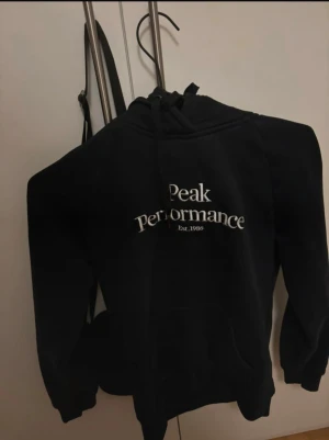 Svart hoodie Peak Performance S - Svart hoodie från Peak Performance med vit logga på bröstet och snörning i huvan. Klassisk kanguruficka framtill och ribbade muddar vid ärmar och nederkant. Perfekt för chill eller när du vill ha en clean look. Köptes för ca 1 år sen men har knappt använts därav säljs den nu köpte den för 600kr 