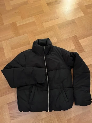 Svart pufferjacka från Jacqueline de Yong XS - Svart pufferjacka från Jacqueline de Yong i storlek XS. Jackan är kortare i modellen, har hög krage och dragkedja framtill. Tillverkad i polyester och perfekt för kyliga dagar. Enkel och stilren design som passar till det mesta.