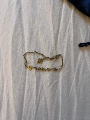 Guldigt armband från Guess - Snyggt armband från Guess i guldfärgad metall med bokstäverna G U E S S och små glittriga stenar. Kedjan har en trekantig Guess-berlock och smidig karbinlås. Perfekt accessoar för att lyfta din stil med lite extra bling.