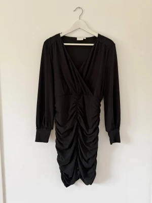 Svart omlottklänning från Vila julbord klänning  - Supersnygg svart kortklänning från Vila med omlott överdel, långa ärmar och draperad kjol. Klänningen har v-ringning och breda muddar vid ärmsluten. Perfekt för dig som gillar en stilren look med coola detaljer.