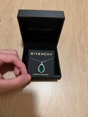 Givenchy halsband med grön Swarovski-kristall - Exklusivt halsband från Givenchy med en droppformad grön Swarovski-kristall omgiven av gnistrande stenar. Kedjan är i silverfärg och smycket levereras i originalask och påse från Givenchy. Perfekt för dig som vill sticka ut med något riktigt lyxigt.