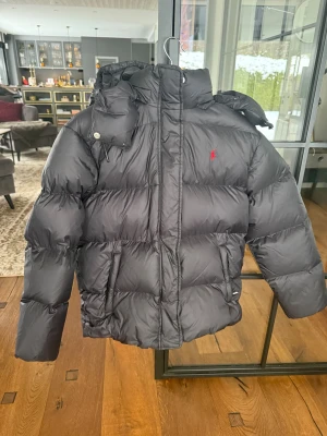 Svart dunjacka Polo Ralph Lauren - Svart dunjacka från Polo Ralph Lauren med avtagbar huva, dragkedja och tryckknappar. Jackan är quiltad och har den klassiska röda Polo-loggan på bröstet. Perfekt för kalla dagar med sin fluffiga och varma känsla.