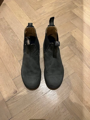 Svarta Chelsea boots från Blundstone - Säljer ett par svarta Chelsea boots från Blundstone med rund tå och platt sula. Svart/grå nubuck. Skorna har elastiska sidopaneler och dragflikar med logga både fram och bak. Tillverkade i slitstark skinn med bekväm innersula. Perfekt för dig som gillar klassisk och stilren look.