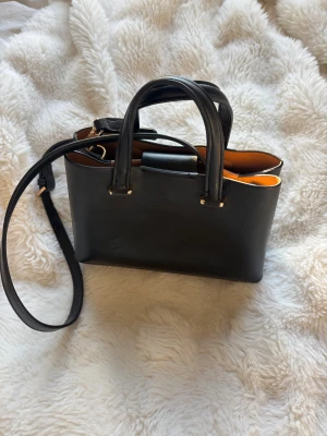 Svart handväska i skinn med guld-detaljer - Stilren svart axelväska i skinn med både handtag och justerbar axelrem. Perfekt för dig som gillar minimalistisk design och med en vardaglig väska som passar till allt. Köpt på Mango för ett år sen och använt fåtal gånger. 