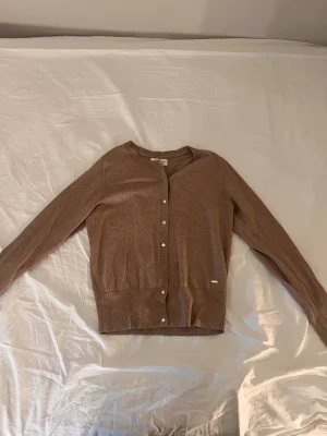 Beige kofta med knappar från Vero Moda - Snygg beige kofta från Vero Moda med rund hals och långa ärmar. Den har knappar framtill och en liten metallbricka vid nederkanten. Mjuk och tunn kvalitet i bomullsmix, perfekt för lager-på-lager under hösten och våren.