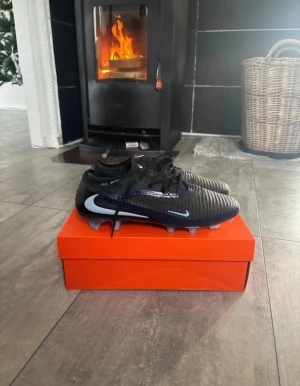 Nike fotbollsskor svarta med blå detaljer - Säljer ett par grymma Nike fotbollsskor i svart med ljusblå och mörkblå detaljer. Skorna har snörning, platt sula med dobbar och en rund tå. Ovandelen är tillverkad i syntetmaterial med texturerad yta för bättre bollkontroll. Perfekt för dig som vill ha stil på planen.
