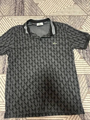 Svart mönstrad piké från Lacoste - Svart pikétröja från Lacoste med grått geometriskt mönster och klassisk krage med vita ränder. Ikoniska gröna krokodilloggan på bröstet. Skön passform och perfekt för dig som gillar stilrena detaljer.