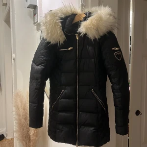 Svart pufferjacka Rockandblue med fuskpäls - Svart pufferjacka från Rockandblue med stor fluffig fuskpäls på huvan, guldfärgade dragkedjor och detaljer. Jackan har quiltade partier på ärmar och sidor, samt Rockandblue-märke på ena ärmen. Perfekt för kalla dagar och riktigt snygg passform.