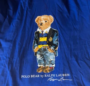 Blå hoodie Polo Bear Ralph Lauren - Blå hoodie från Ralph Lauren med Polo Bear-tryck på bröstet. Tröjan har långa ärmar, huva och är gjord i mjuk bomull. Trycket visar en nalle i preppy stil med jeans och kavaj. Perfekt för dig som gillar klassisk grisch med en twist.