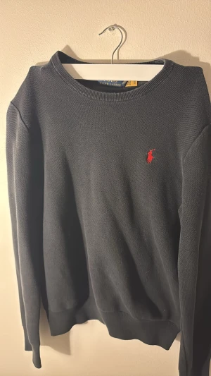 Svart stickad tröja Polo Ralph Lauren - Svart stickad tröja från Polo Ralph Lauren i storlek M. Klassisk rund hals och långärmad modell med röd broderad logga på bröstet. Tröjan har ribbade muddar vid ärmslut och nederkant, perfekt för en clean och stilren look.