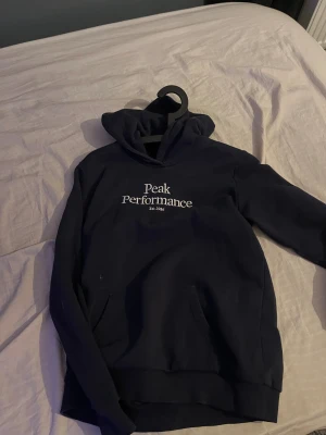 Mörkblå hoodie Peak Performance 170 - Snygg mörkblå hoodie från Peak Performance med vit logga på bröstet. Tröjan har huva, magficka och långa ärmar. Perfekt för chill dagar och när du vill ha en clean look. Mjuk insida i bomullsmix som gör den skön att bära.
