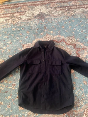 Mörkblå overshirt från H&M - Säljer en stilren mörkblå overshirt från H&M med klassisk krage och två stora bröstfickor med lock. Jackan har knäppning framtill och är tillverkad i ett mjukt material som passar perfekt för lager-på-lager. Enkel och clean look som funkar till det mesta.              MÖTS I STOCKHOLM🙃
