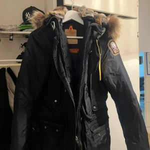 Svart Parajumpers parkas med pälskrage - Säljer en svart Parajumpers parkas med ikonisk patch på ärmen, gul detalj vid halsen och stor huva med fluffig pälskant. Jackan har flera fickor, dragkedja och knappar framtill. Perfekt för kalla dagar och riktigt snygg streetstil.