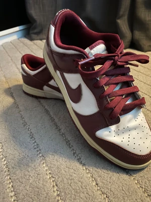 Nike Dunk Low Bordeaux/Vit - Nike Dunk Low sneakers i färgen bordeaux och vit. Klassisk siluett med lågt skaft, snörning och perforerad tåbox. Ovandel i skinn med textilfoder och matchande bordeauxfärgade skosnören. Snygg kontrast mellan de vita och vinröda panelerna. Perfekta för dig som gillar streetwear.