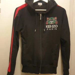 Svart hoodie från Kenzo - Snygg svart hoodie från Kenzo med färgglatt tigertryck och texten 'Kenzo Paris' på både fram- och baksidan. Hoodien har en dragkedja och röda ränder längs ärmarna. Perfekt för en cool och avslappnad stil. Storlek M men känns mer som en S
