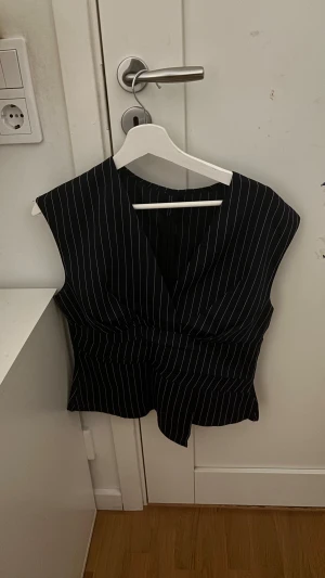 Mörkblå randig omlott blus - Säljer en mörkblå blus med vita ränder och omlott-design. Toppen har breda axlar och en snygg v-ringning som ger en cool siluett. Perfekt att styla med jeans eller kjol för en trendig look. Materialet känns mjukt och följsamt mot kroppen.