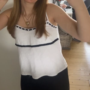 Vit linnetopp med svarta detaljer - Säljer en vit linnetopp med svarta detaljer och tunna axelband. Toppen har en dekorativ svart kant upptill och ett svart band som går horisontellt över bröstet. Materialet känns lätt och luftigt, perfekt för varma dagar. Köpte för 500kr för några månader sedan