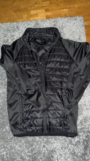 Svart pufferjacka från Acode, M - Svart pufferjacka från Acode i storlek M. Jackan har quiltade paneler framtill och baktill, släta svarta ärmar och dragkedja hela vägen. Snygg, sportig look med två sidofickor med dragkedja. Perfekt för dig som gillar stilren och funktionell design.