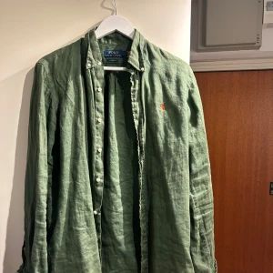 Grön linneskjorta från Polo Ralph Lauren - Snygg grön skjorta från Polo Ralph Lauren i linne med custom fit. Klassisk krage, vita knappar och lång ärm. Diskret orange logga broderad på bröstet. Perfekt för dig som gillar stilrena och bekväma plagg med lite edge.