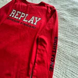 Rullat  långärmad tröja från Replay - Snygg röd långärmad tröja från Replay med tryck 'REPLAY SUPERIOR STANDARD' på ryggen och 'EIGHTYONE' på ena ärmen. Tröjan är i mjuk bomull och har en klassisk rund halsringning. Perfekt för dig som gillar streetwear vibes.