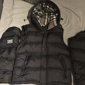 Svart pufferjacka från Burberry och väst - Säljer en svart pufferjacka från Burberry med avtagbara ärmar så den kan användas som väst. Jackan har huva med klassiskt rutigt foder i grått, vitt, svart och rött. Stora fickor med dragkedja och tydlig Burberry-patch på ärmen. Perfekt för kalla dagar.