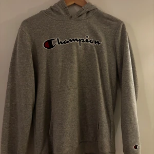 Grå hoodie från Champion XL pojke  - Snygg grå hoodie från Champion i storlek XL i barnstorlek med klassisk logga på bröstet och ärmen. Tröjan har huva och en stor magficka framtill. Perfekt för en chill och sportig stil. Tillverkad i mjuk bomullsmix för extra komfort.