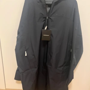 Svart regnjacka från Peak Performance - Svart regnjacka från Peak Performance med huva och hel dragkedja framtill. Jackan har minimalistisk design, diskret logga på bröstet och praktiska fickor med dragkedja. Perfekt för blöta dagar och enkel att matcha med allt.