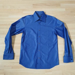 Blå skjorta från Merkur Linnefabriken - Snygg blå skjorta från Merkur Linnefabriken 1919 i storlek M. Skjortan har klassisk krage, långa ärmar med dubbla manschetter och svarta knappar. Tillverkad i bomull för en skön känsla och stilren look.