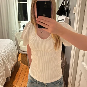 Vit ribbad topp med puffärm och knyt - Superfin vit ribbad topp med korta puffärmar och v-ringning både fram och bak. Toppen har en söt knyt-detalj i ryggen och är figurnära i modellen. Prislappen sitter kvar och har aldrig använts. 