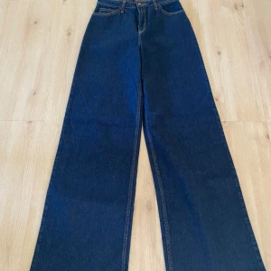 Mörkblå wide jeans från Lee - Snygga mörkblå jeans från Lee med klassisk femficksdesign och raka, vida ben. Hög midja och tydliga kontrastsömmar ger en retrovibe. Jeansen är tillverkade i robust denim och har Lee-logga på bakfickan och knappen framtill.