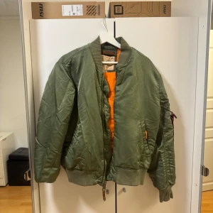 Grön bomberjacka från Alpha Industries - Säljer en klassisk grön bomberjacka från Alpha Industries med orange foder. Jackan har dragkedja framtill, ribbade muddar och är långärmad. Perfekt för dig som vill ha en streetig och tidlös look.