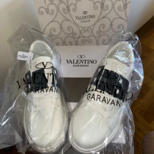 Valentino Garavani - Snygga vita sneakers från Valentino Garavani med svart band och diskreta nitar på sidorna. Skorna har rund tå, platt sula och är tillverkade i skinn. Klassisk och stilren design med logga på plösen och sulan. Levereras med originalkartong och dustbag.