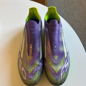 Adidas F50 lila fotbollsskor - Säljer ett par Adidas F50 fotbollsskor i lila och neongult med snyggt grafiskt mönster. Skorna har strumpliknande passform utan snörning och är tillverkade i syntetmaterial. Sulan har fasta dobbar för konstgräs. Medföljer matchande sko påse