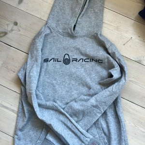 Grå hoodie från Sail Racing, strl 12 år  - Snygg grå hoodie från Sail Racing med svart logga på bröstet. Klassisk modell med huva och känguruficka framtill. Perfekt för dig som gillar sportig och avslappnad stil.