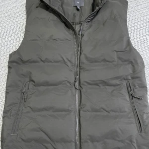 Mörkgrön dunväst från H&M - En stilren mörkgrön dunväst från H&M med hög krage och två fickor med dragkedja. Västen har quiltad design och hel dragkedja framtill. Västen är använd använd 1-3 gånger och inga täcken på användning eller märken/fel. Storlek S. Västen är dessutom vattenavvisande och vindtät. 