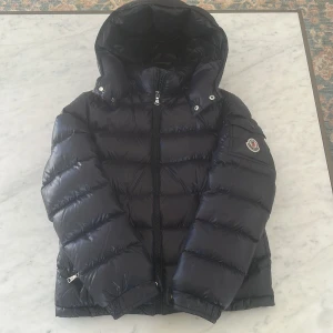 Marinblå Moncler pufferjacka med huva - Säljer en marinblå pufferjacka från Moncler med stor huva och klassisk logga på ärmen. Jackan har dragkedja framtill, fickor och quiltad design. Perfekt för kalla dagar och riktigt snygg streetstil.