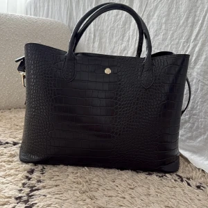 Svart handväska med crocomö-mönster - Svart handväska i croco-mönstrad skinnimitation med två handtag, reglerbar och avtagbar axelrem och magnetlås. Väskan har en rymlig insida med mittenfack med guldfärgad dragkedja. På ena sidan om dragkedjefacket finns små fack - ett för kort och två för mobiltelefon. Plats för laptop. 