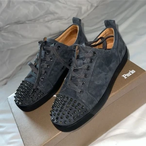 Christian Louboutin svarta mocka sneakers - Snygga svarta sneakers från Christian Louboutin i mocka med ikoniska röda sulor och svarta nitar på tån. Skorna har snörning och en cool, edgy vibe som sticker ut. Perfekta för dig som vill ha något unikt och exklusivt i garderoben.